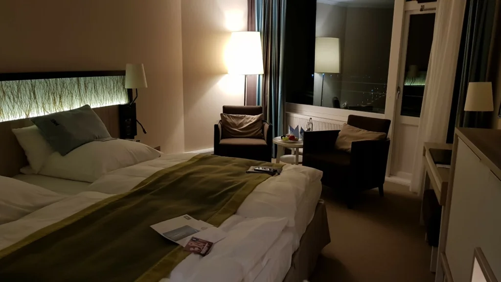Elegantes Hotelzimmer im warmen Licht: Ein großes Doppelbett mit grüner Bettdecke und weißem Kopfkissen, darüber eine stilvolle Beleuchtung mit einem Muster, das an Zweige erinnert. Links im Raum steht ein komfortabler Ledersessel neben einer Tischlampe, die eine sanfte Beleuchtung verbreitet. Rechts führt eine Balkontür hinaus, durch die man einen Blick auf die nächtliche Stadtlandschaft erhaschen kann. Auf dem Bett und dem Beistelltisch befinden sich Informationsbroschüren und ein kleines Obstteller.