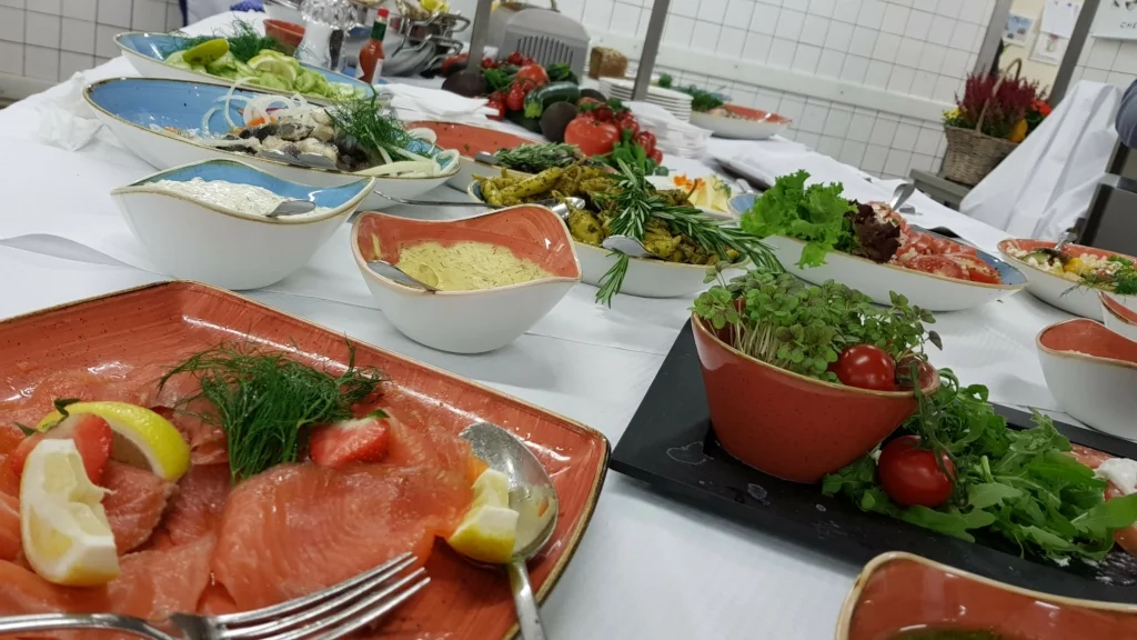 Vielfältiges Buffet in einer professionellen Küche: Auf Vordergrund ein orangefarbener Teller mit dünn geschnittenem Lachs, garniert mit Zitronenscheiben und Dill. Dahinter verschiedene Schalen gefüllt mit cremigem Hummus, grünen Oliven, eingelegtem Gemüse und frischen Salaten. Weiter hinten sieht man bunte Gemüsesorten und dekorative Kräutertöpfe, alles ansprechend präsentiert auf einer weißen Tischdecke.