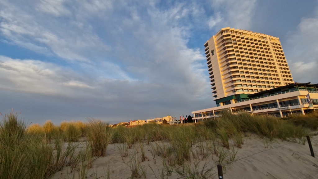 Hotelgebäude am Strand mit hohem Wolkenkratzer, umgeben von Dünen und Gras, unter einem bewölkten Himmel bei Sonnenuntergang.